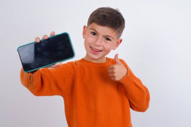 Beyaz arka planda dikilen örgü turuncu süveter giyen beyaz çocuk. Hücrede el ele tutuşmuş, siyah başparmağını kaldırıyor.