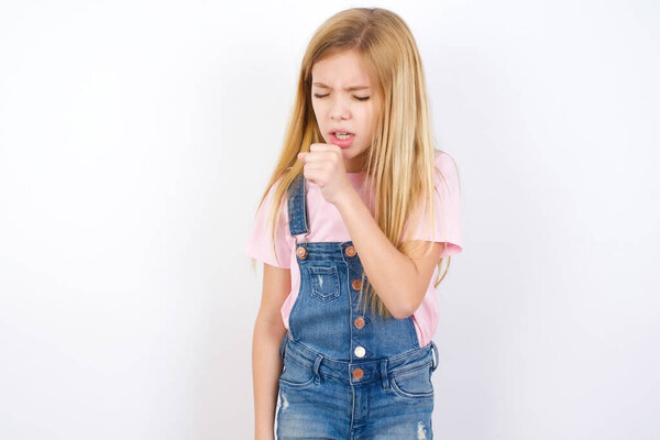 beautiful caucasian little girl wearing denim jeans overall over white background feeling unwell and coughing as symptom for cold or bronchitis. Healthcare concept.