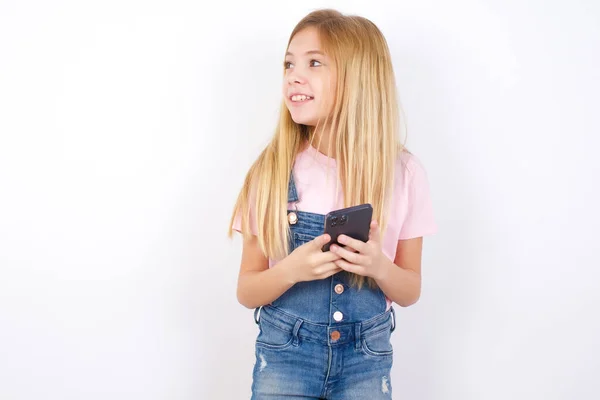 白い背景の上にデニムジーンズを身に着けている美しい白人の女の子は スマートフォンを保持し 空白のコピースペースを横に見て — ストック写真