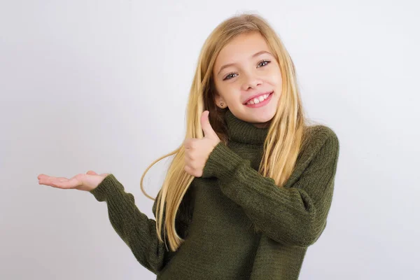 白い背景に緑のニットセーターを着て白人の子供の女の子ヤシの手を表示し 親指でOkジェスチャーを行います 笑顔幸せと陽気な — ストック写真
