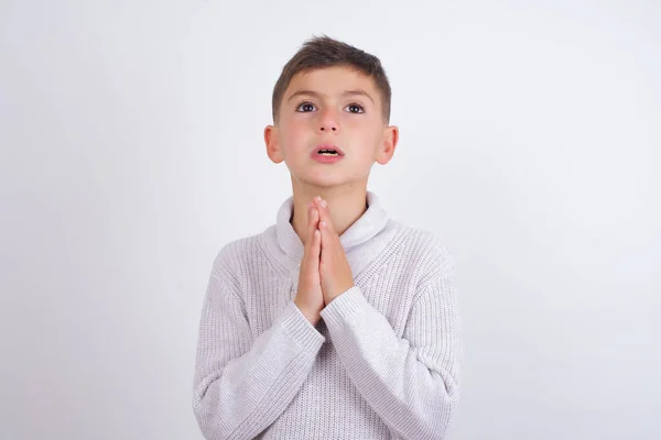 白地の上に立つニットセーターを着た白人の少年は 非常に感情的で心配している顔に希望の表情と一緒に手で物乞いと祈りをしています — ストック写真