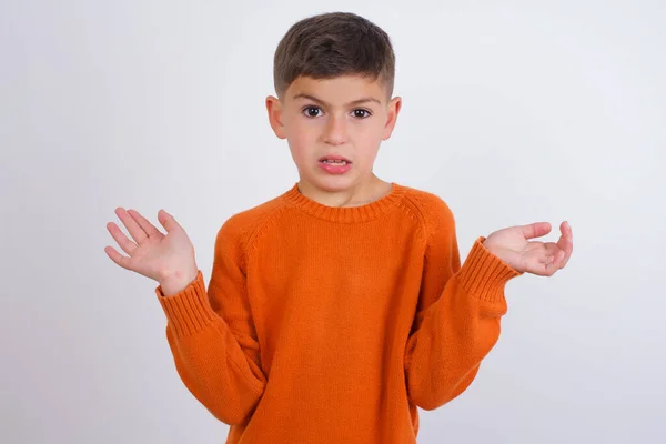 怒气冲冲的白人小男孩穿着针织的橙色毛衣站在白色的背景图片前 困惑得皱着眉头 满脸不悦 — 图库照片