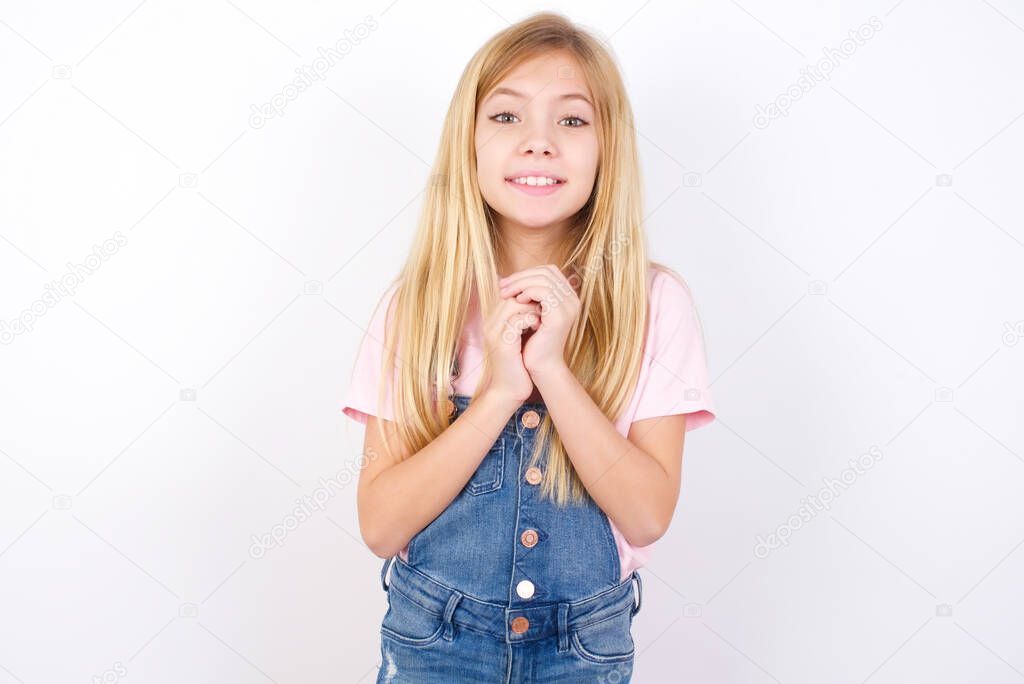 Positive beautiful caucasian little girl wearing denim jeans overall over white background smiles happily, glad to receive pleasant news from interlocutor, keeps hands together. People emotions concept.