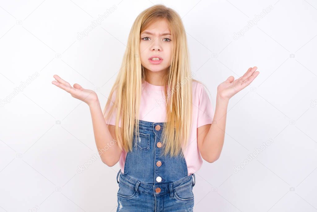 Indignant beautiful caucasian little girl wearing denim jeans overall over white background gestures in bewilderment, frowns face with dissatisfaction.