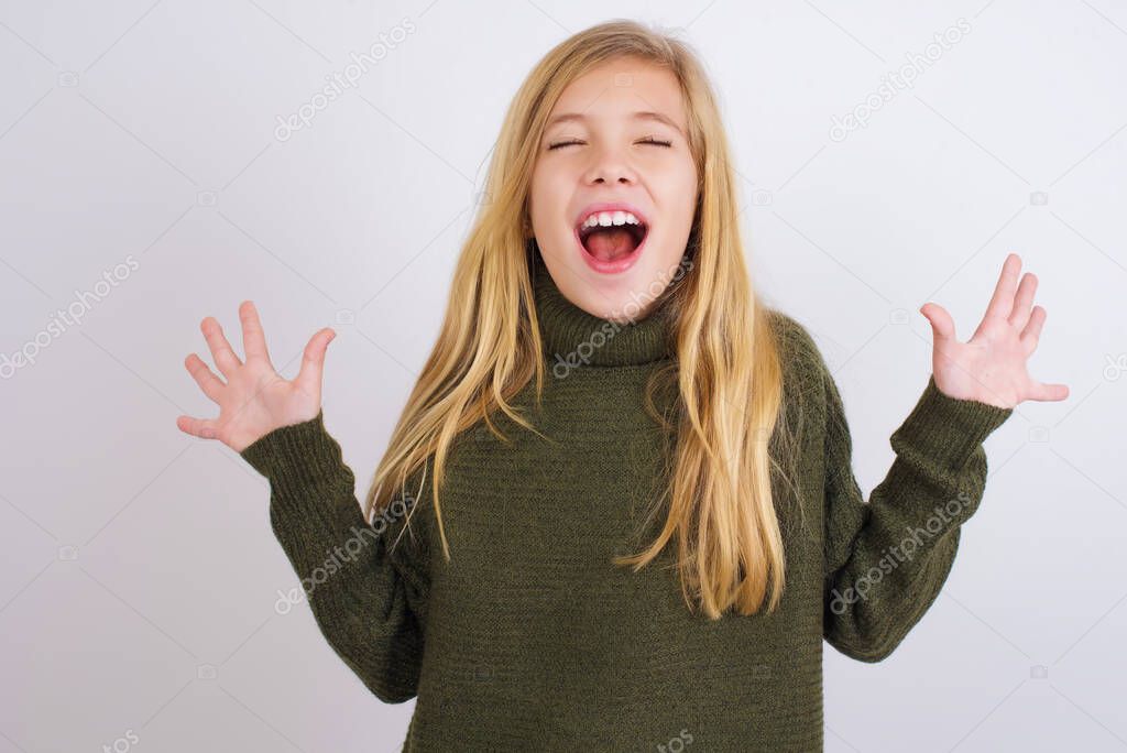 Emotive Caucasian kid girl wearing green knitted sweater against white background laughs loudly, hears funny joke or story, raises palms with satisfaction, being overjoyed amused by friend