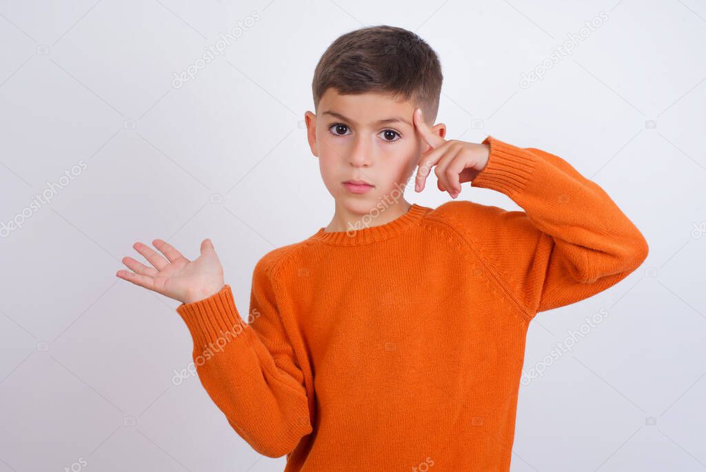 Caucasian kid boy wearing knitted orange sweater standing over white background confused and annoyed with open palm showing copy space and pointing finger to forehead. Think about it.
