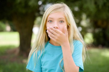 Emotional Caucasian little kid girl wearing blue T-shirt standing outdoors gasps from astonishment, covers opened mouth with palm, looks shocked at camera. Reaction concept clipart