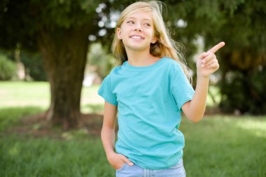 Mavi tişört giyen, gülümseyen beyaz bir kız çocuğu. Açık havada duruyor. Boş bir parmağın en düşük fiyatları gösterdiğini gösteriyor.