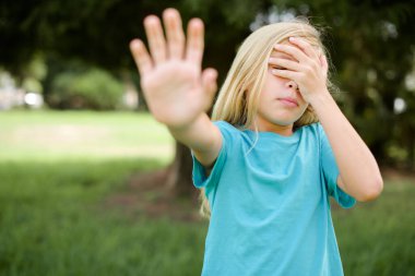 Açık havada duran mavi tişörtlü, beyaz bir kız çocuğu. Avuç içiyle gözlerini kapatıyor ve jest yapıyor. Herkesten saklanıyor..