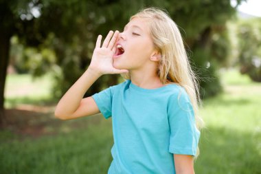 Caucasian little kid girl wearing blue T-shirt standing outdoors shouting and screaming loud to side with hand on mouth. Communication concept. clipart