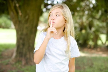 Beyaz tişörtlü, açık havada duran beyaz çocuk jesti parmakları dudaklara işaret ediyor. Sırrımızı paylaşmamamızı istiyor.