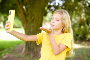 Açık havada duran sarı tişörtlü güzel beyaz kız akıllı telefonun kamerasına hava öpücüğü veriyor ve selfie çekiyor, internetten Mwah 'ı gönderiyor..