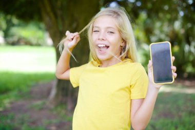 Güzel, güzel, beyaz, sarı tişörtlü bir kız çocuğunun fotoğrafı. Açık havada duruyor. Telefon ekranı saç kuyruğu tutuyor.