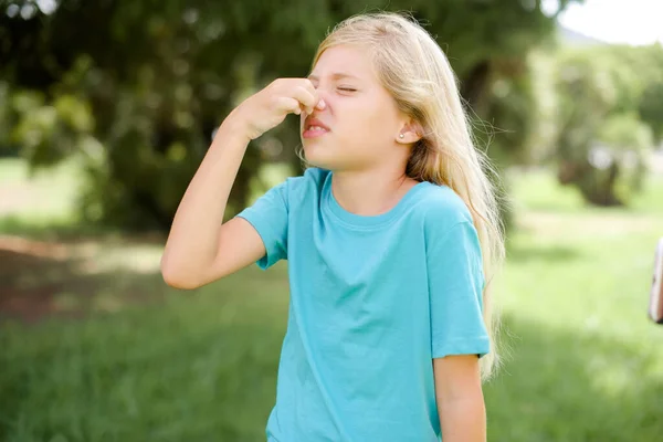 Caucasian little kid girl wearing blue T-shirt standing outdoors smelling something stinky and disgusting, intolerable smell, holding breath with fingers on nose. Bad smell