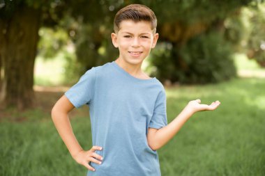 Güzel beyaz çocuk mavi tişört giymiş dışarıda mutlu ve neşeli duruyor, sana gülümsüyor ve hoş geldin diyor, seni dostça bir jestle içeri davet ediyor.