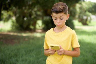 Açık havada duran sarı tişörtlü güzel beyaz çocuk akıllı telefonlardan sosyal medya haberleri veya önemli e-postalar okuyor.