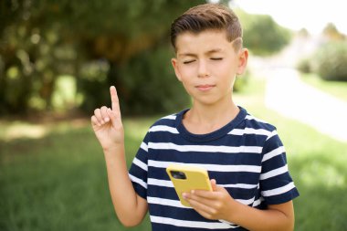 Gülümseyen beyaz çocuk çizgili tişört giyiyor açık havada boş bir yeri işaret ediyor telefonu bir elinde tutuyor.