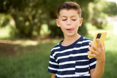 Güzel, güzel, beyaz, çizgili tişört giyen bir çocuğun fotoğrafı. Açık havada duruyor. Telefon ekranı saç kuyruğu tutuyor.