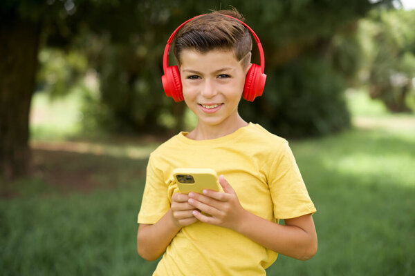 Positive beautiful Caucasian little kid boy wearing yellow T-shirt standing outdoors holds modern cell phone connected to headphones, clenches fist from good emotions, exclaims with joy,