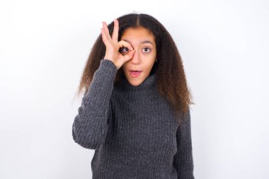 Gri balıkçı yaka giyen güzel bir genç kız beyaz arka planda dikiliyor. Şok olmuş bir jest yapıyor. İnançsız ifade.