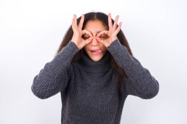 Gri balıkçı yaka giyen güzel bir genç kız beyaz arka planda dikiliyor. Dürbün gibi güzel hareketler yapıyor, gözleri parmaklarından bakıyor. Çılgın ifade.