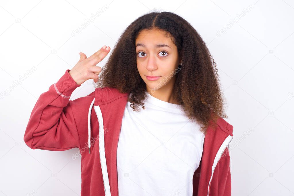Unhappy beautiful teen girl wearing casual clothes standing against white background makes suicide gesture and imitates gun with hand, curves lips, keeps two fingers on temple