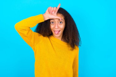 Sarı süveter giyen güzel bir genç kız mavi arka plan jestlerine karşı ayakta duruyor. Parmağını alnına koyuyor. Ezik jestler yapıyor.