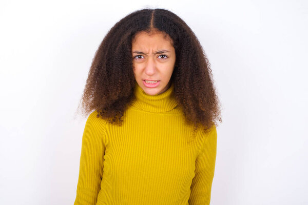 красивая девочка-подросток в желтой водолазке, стоящая на белом фоне держит зубы сжатыми, хмурится лицом в неудовлетворенности, раздражается из-за многих обязанностей.