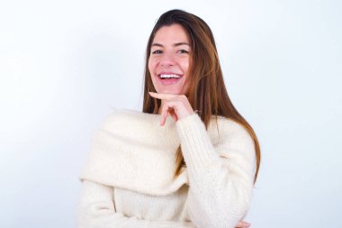 İyimser, beyaz arka plan giyen genç bir kadın ellerini çapraz tutuyor ve elleri çenesinin altında tutuyor. Kameraya zevkle bakıyor. Mutlu duygu konsepti.