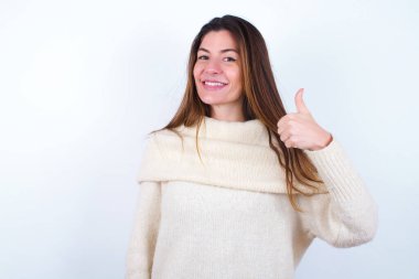 Beyaz süveter giyen genç bir kadın elleriyle mutlu bir jest yapıyor. Kameraya bakıp başarıyı gösteren ifade onaylanıyor.