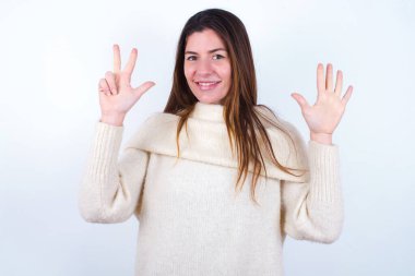 Beyaz süveter giyen genç bir kadın 8 numaralı parmaklarıyla gülümseyerek mutlu ve kendinden emin bir şekilde öne çıkıyor..