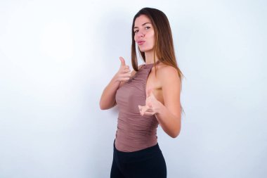 Spor kıyafetleri giyen neşeli genç bir kadın beyaz arka plan göz kırpıyor ve işaret parmaklarıyla kameraya işaret ediyor, birini seçiyor, parmak tabancası yapıyor..