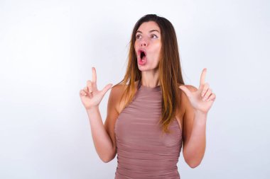 Spor kıyafetleri giyen genç bir kadın şaşırmış ve şaşırmış bir şekilde yukarı bakıyor ve parmaklarıyla işaret ediyor ve kollarını kaldırıyor..