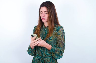 Beyaz arka plan üzerine çiçekli elbise giyen heyecanlı beyaz kadın göz kırpıyor ve akıllı telefon kullanımı sosyal ağ haberlerini okuyor.