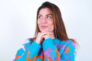 Curious Young caucasian woman wearing vintage colorful sweater over white background keeps hands under chin bites lips and looks with interest aside.