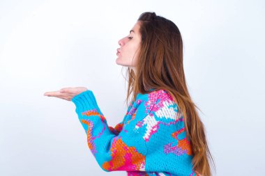 Profile side view view portrait of attractive Young caucasian woman wearing vintage colorful sweater over white background sending air kiss