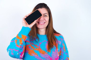 Young caucasian woman wearing vintage colorful sweater over white background holding modern smartphone covering one eye while smiling
