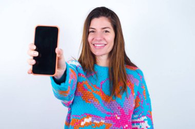 Charming adorable Young caucasian woman wearing vintage colorful sweater over white background holding modern device, showing black screen smartphone