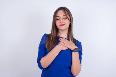 Beyaz arkası üzerine mavi elbise giyen genç Arap kadın gözlerini kapar ve ellerini kalbine yakın göğsüne koyar, samimi duygularını ifade eder, iyi kalpli ve dürüst davranır. Vücut dili ve gerçek duygular kavramı.