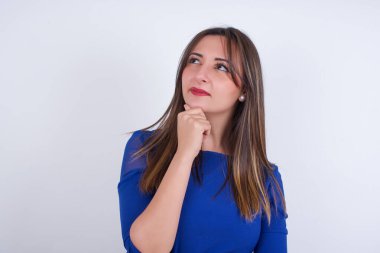 Düşünceli, genç bir Arap kadının beyaz arkası üzerine mavi elbise giyip elini çenesinin altında tutması, düşünceli bir şekilde yukarı bakması..