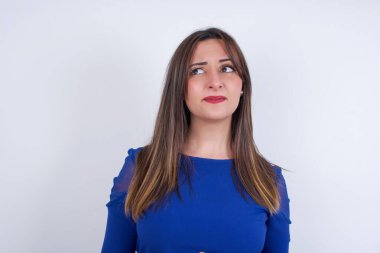 Şaşırmış genç Arap kadın beyaz sırt üstü mavi elbise giyiyor, dudaklarını kıvırıyor ve endişeli bakıyor, önünde korkunç bir şey görüyor..