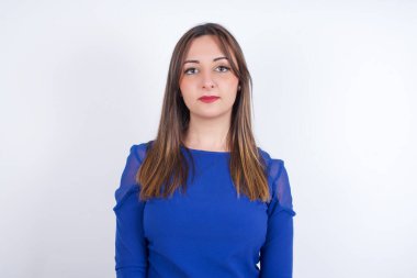 Mavi elbiseli, beyaz sırtlı neşeli genç Arap kadın kameraya bakıyor, bir şey düşünüyor. İki kolunu da indir, tarafsız yüz ifadesi..