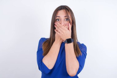 Kötü bir şey görüp duyduktan sonra avuçlarını iki avuçla kapatan, beyaz arka planda mavi elbise giyen üzgün genç Arap kadın..