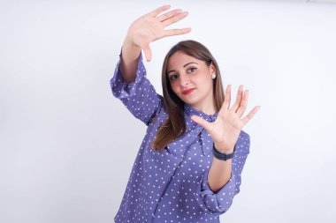 Beyaz sırt çantasına benekli giysiler giymiş gülümseyen Arap kadın portresi kameraya bakıyor ve işaret eden parmak çerçevesine bakıyor. Yaratıcılık ve fotoğrafçılık kavramı.