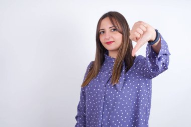 Hoşnutsuz genç Arap kadın beyaz arka plan üzerinde benekli giysiler giyiyor. Onaylamıyor, baş parmağını aşağıda tutuyor, hoşnutsuzluğunu ifade ediyor, hoşnutsuzluğunu ifade ediyor. Olumsuz duygular.