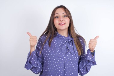 İyimser genç Arap kadın beyaz sırt çantasına benekli giysiler giyip olumlu duygularla baş parmağını kaldırıyor. Kalite ve tavsiye kavramı.