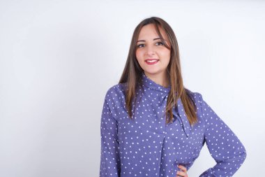 Beyaz sırt çantasının üzerine benekli giysiler giyen neşeli Arap kadının stüdyo fotoğrafı elini kalçasında tutuyor, geniş bir şekilde gülümsüyor.