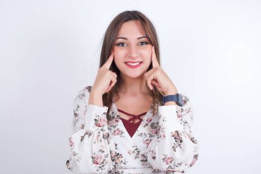 Güçlü, sağlıklı, beyaz dişler. Beyaz arka planda çiçekli tişört giyen mutlu Arap kadının portresi. Parlayan gülümsemesi kusursuz beyaz dişleri işaret ediyor..