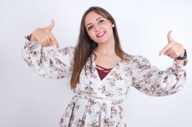 Beni seç! Kendine güvenen, kendine güvenen, karizmatik, beyaz arka planda çiçekli tişört giyen genç bir Arap kadın. Kendini büyük bir gülümseme ve vücuduna işaret eden bir rol istiyormuş gibi tanıtıyor..