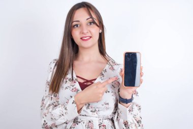 Gülümseyen Arap kadın, beyaz arka bahçeye çiçek desenli gömlek giyip boş telefon ekranını işaret ediyor. Reklam ve iletişim konsepti.